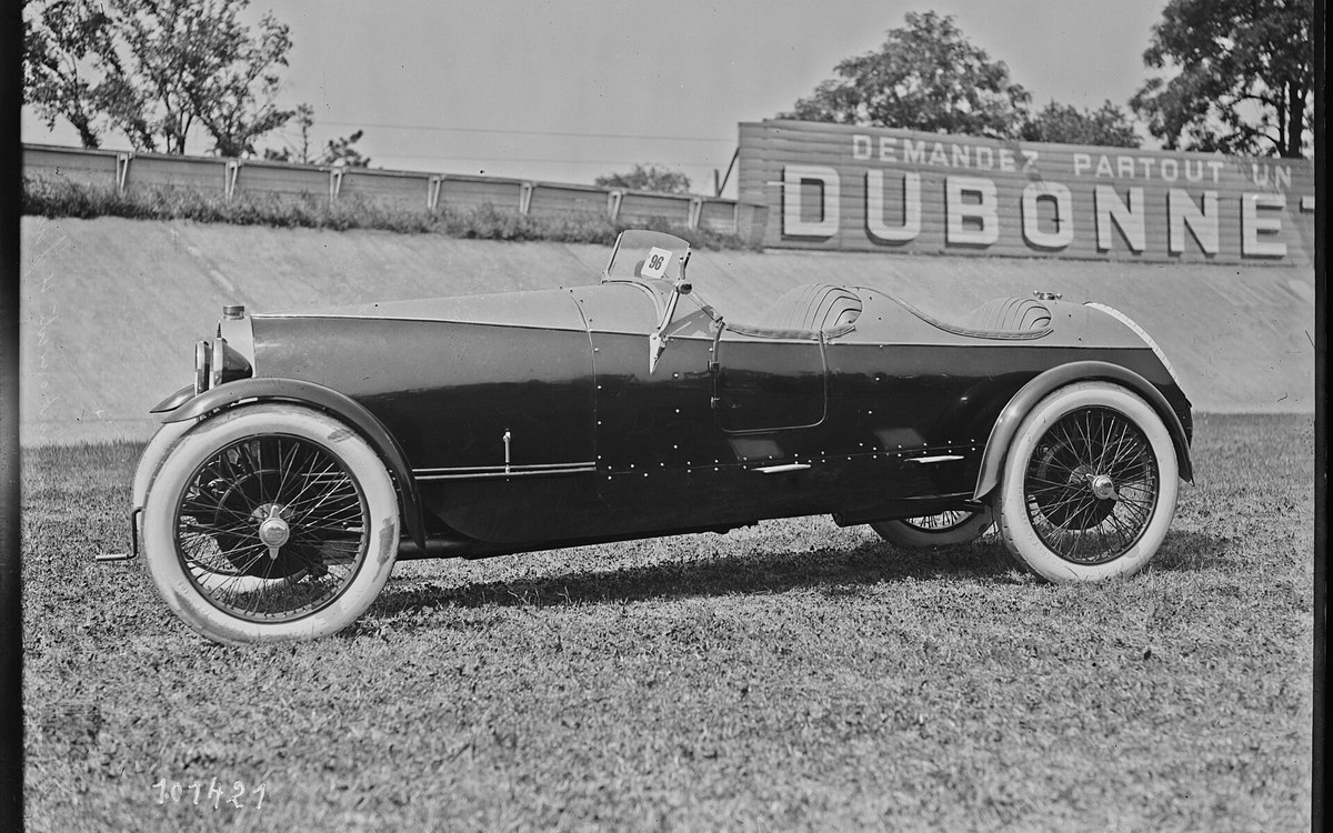 Automóvil de carreras de 1925 visto de perfil sobre la hierba de un circuito, con carrocería alargada, dorsal en el parabrisas y neumáticos de banda blanca.