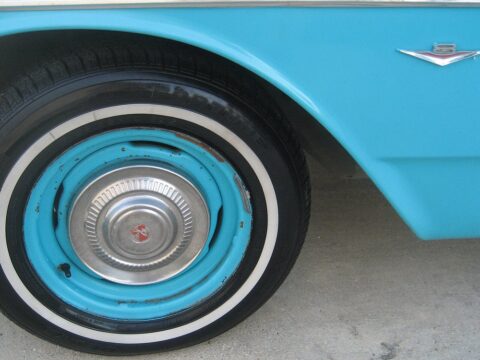 Rueda trasera de un automóvil clásico turquesa, con tapacubos cromado, neumático de banda blanca y el emblema V8 sobre la aleta.