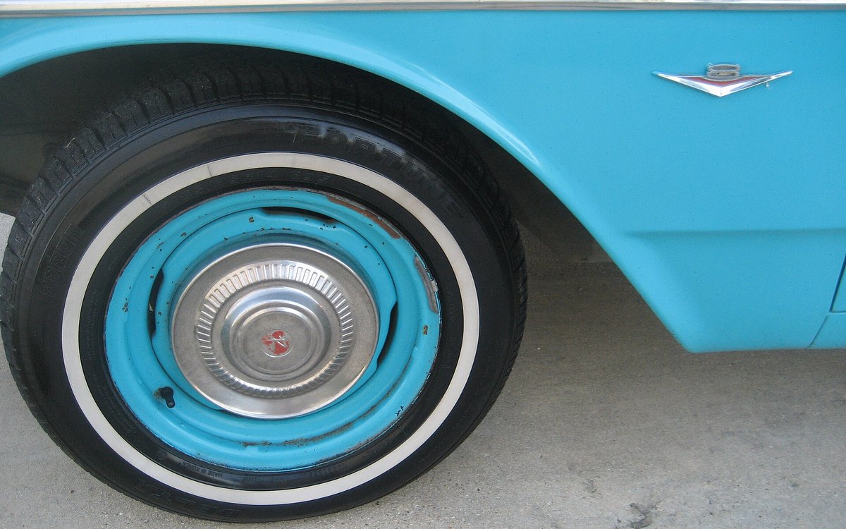 Rueda trasera de un automóvil clásico turquesa, con tapacubos cromado, neumático de banda blanca y el emblema V8 sobre la aleta.