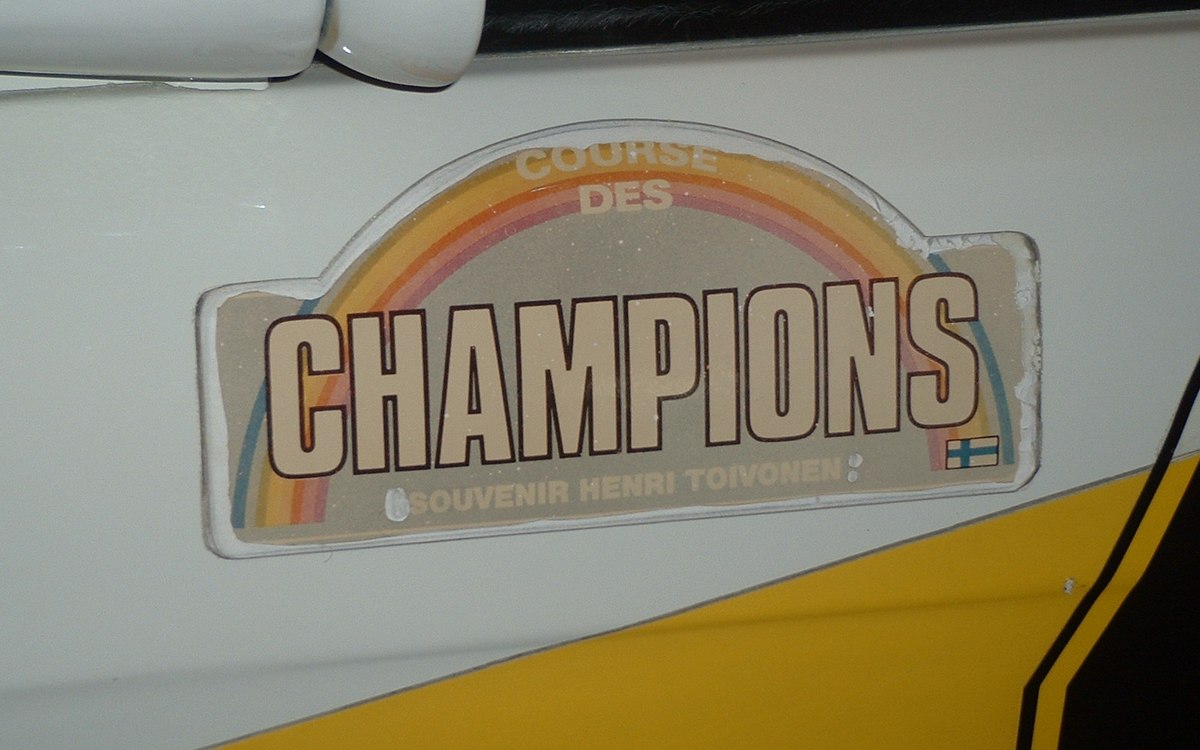 Placa conmemorativa pegada en el lateral blanco y amarillo de un coche de rally de Grupo B, con el texto «Course des Champions — Souvenir Henri Toivonen» y una bandera de Finlandia.