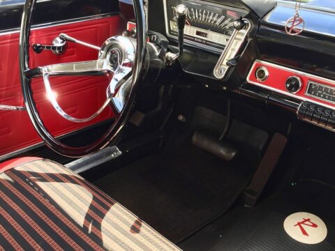 Interior de un automóvil clásico de 1963: volante de dos radios, salpicadero con relojes cromados, tapicería roja y negra a rayas y radio de la época.