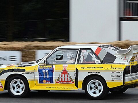 Coche de rally del Grupo B con decoración blanca y amarilla y un gran alerón trasero, rodando de perfil sobre asfalto en una exhibición de vehículos históricos.