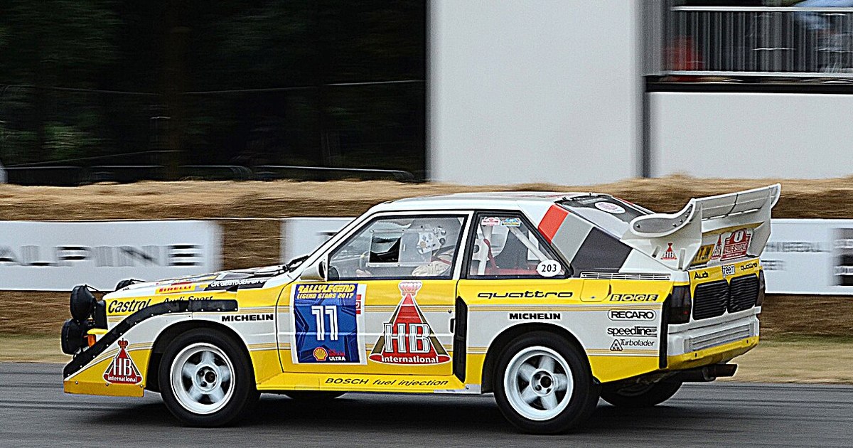 Coche de rally del Grupo B con decoración blanca y amarilla y un gran alerón trasero, rodando de perfil sobre asfalto en una exhibición de vehículos históricos.
