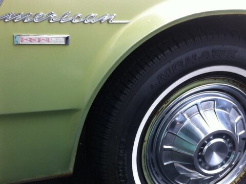 Aleta delantera de un Rambler American de 1968 en verde oliva, con el emblema cromado del modelo y una rueda de tapacubos brillante con neumático de banda blanca.