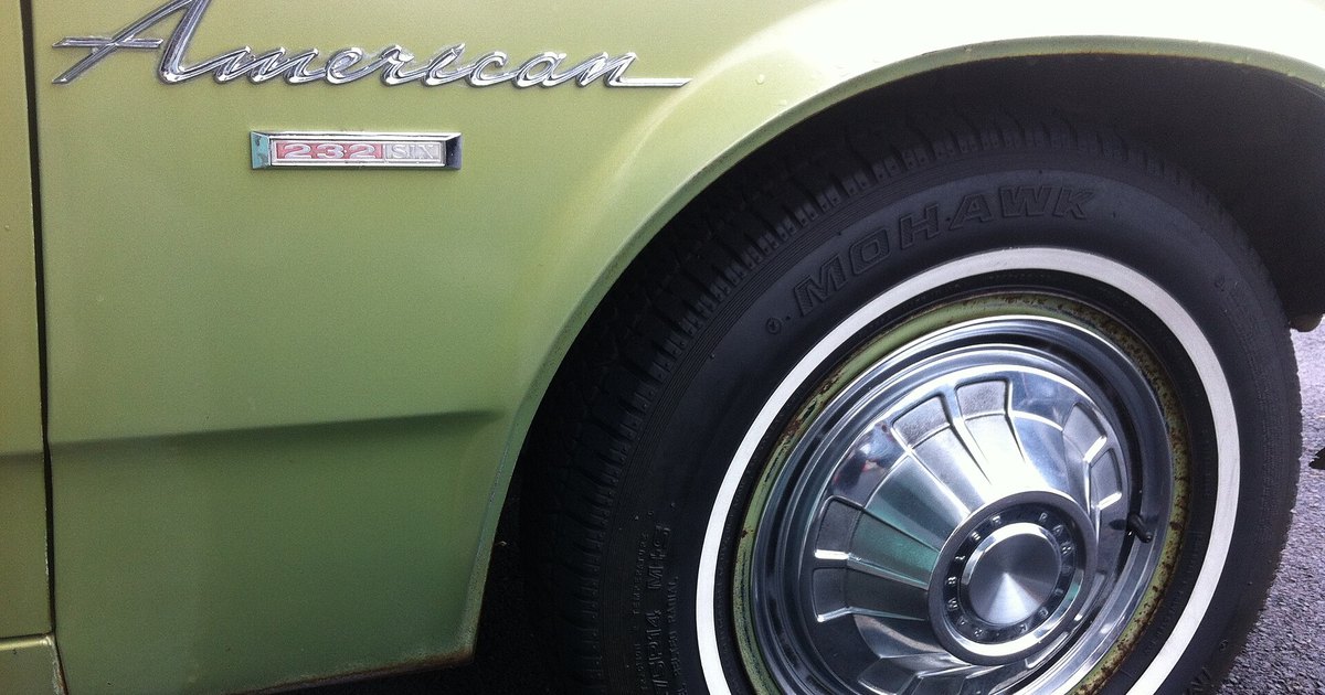 Aleta delantera de un Rambler American de 1968 en verde oliva, con el emblema cromado del modelo y una rueda de tapacubos brillante con neumático de banda blanca.
