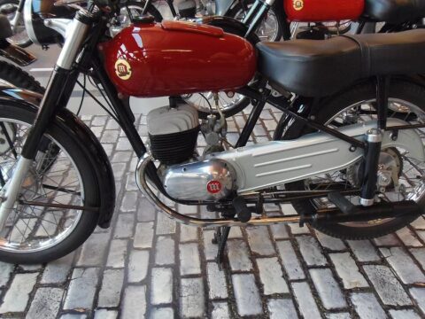 Motocicleta Montesa Comando 150 de 1961, con depósito rojo y motor de dos tiempos, expuesta sobre adoquines en un museo, con más motos clásicas al fondo.