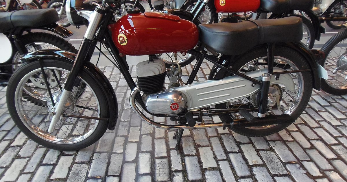 Motocicleta Montesa Comando 150 de 1961, con depósito rojo y motor de dos tiempos, expuesta sobre adoquines en un museo, con más motos clásicas al fondo.